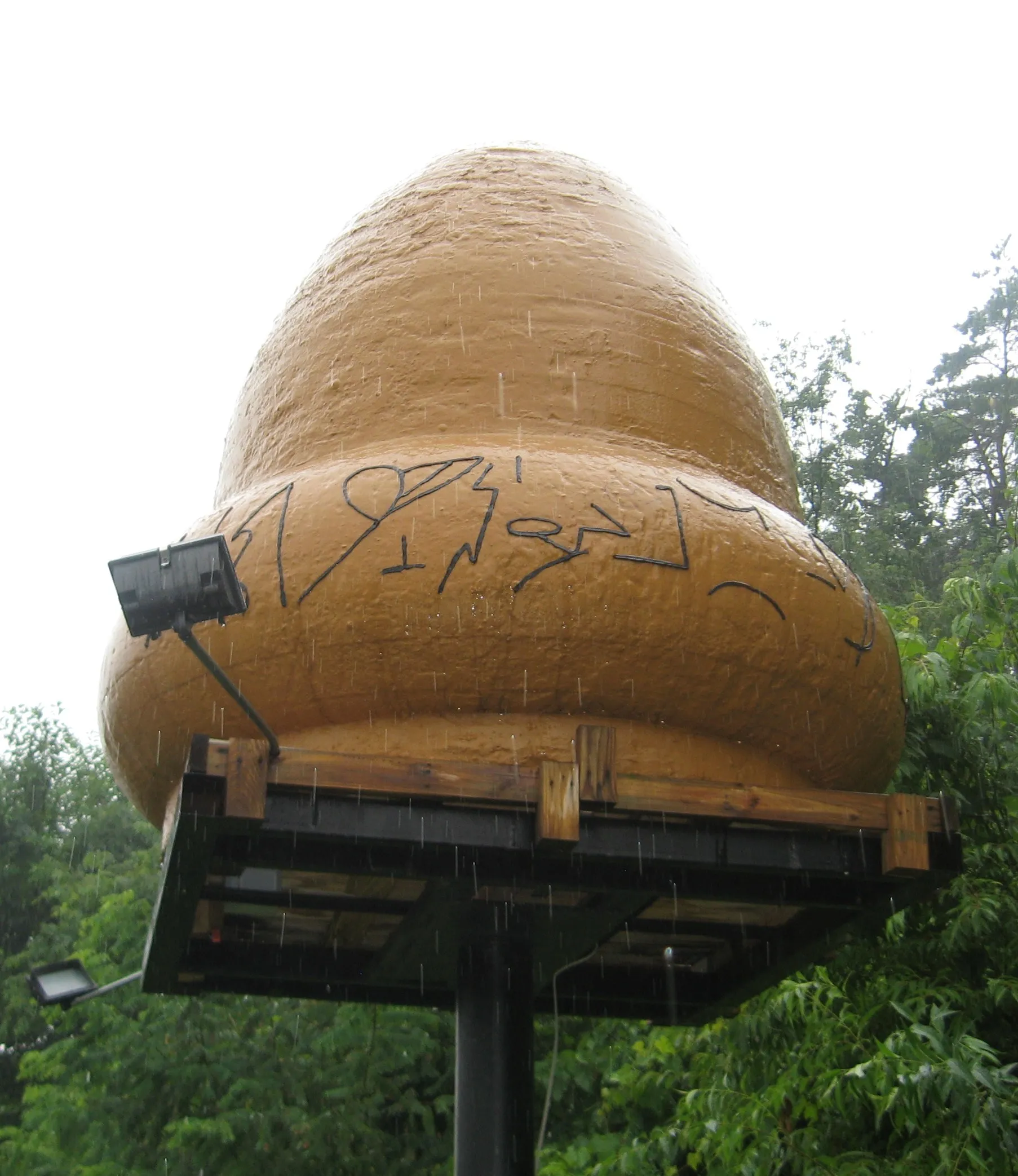 Acorn-shaped metal sculpture with hieroglyphic markings around its base, displayed outdoors next to a small building.