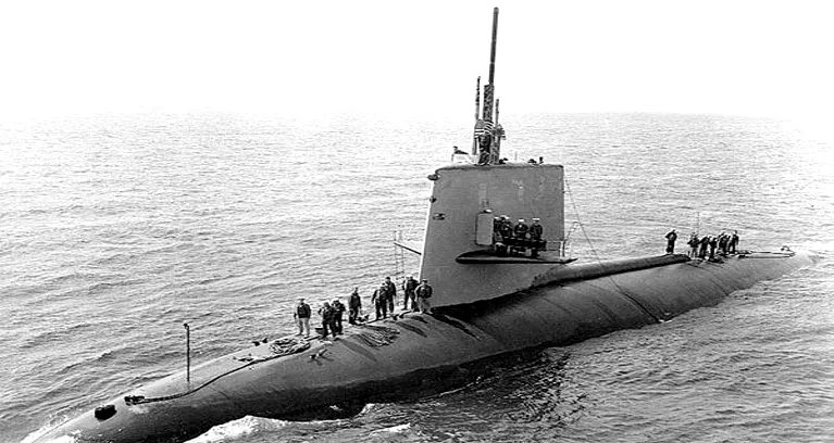 Black-and-white photograph of USS Scorpion, a nuclear-powered submarine, on the surface entering port with crew on deck.